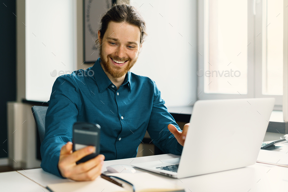 Smiling young man in casual wear looking at smartphone screen at workplace - Stock Photo - Images