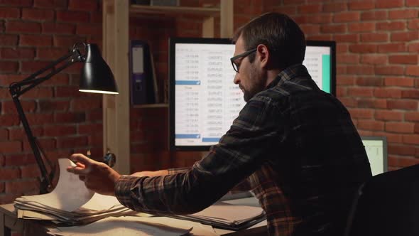 Back View of Caucasian Male in Glasses Sitting in Front of Monitr and Foraging Among Papersin alt