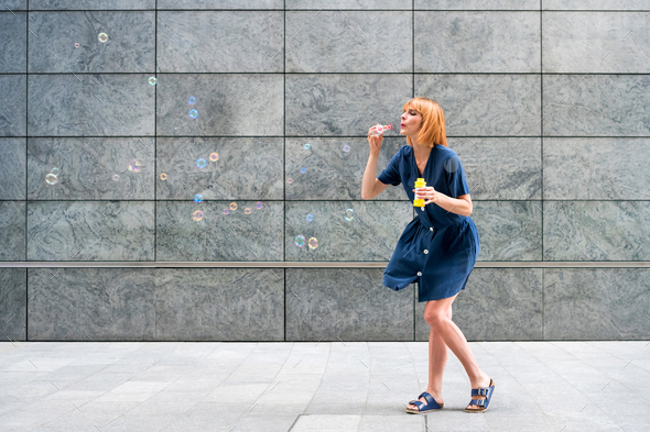 Carefree redhead woman celebrating blowing soap bubbles in town - Stock Photo - Images