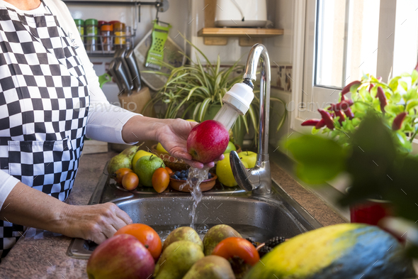 Hands of a senior woman in the kitchen washing a red apple under the running water. - Stock Photo - Images