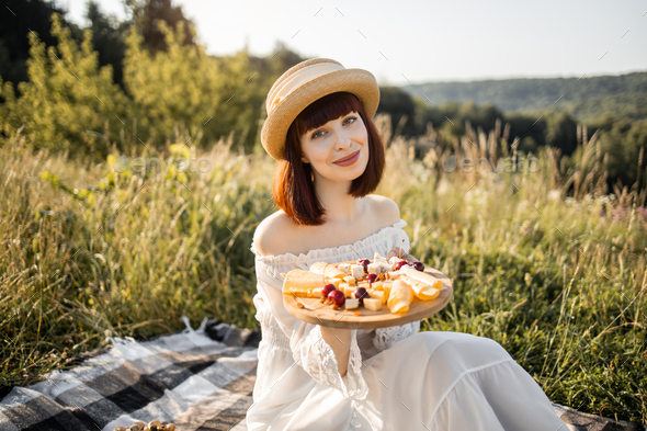 lady in dress and hat, sitting on the blanket with wooden plate with cheese and sweet cherries