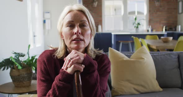 Thoughtful senior caucasian woman in living room sitting on sofa, holding walking cane alt