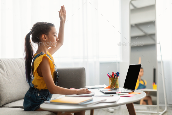 Girl sitting at table, using laptop, raising hand for answer