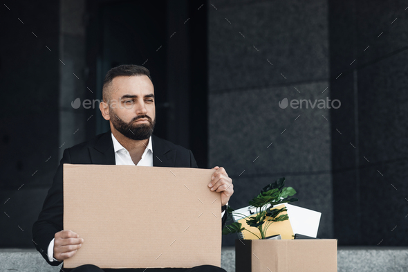Upset fired male employee sitting outside and holding empty poster with box of personal belongings