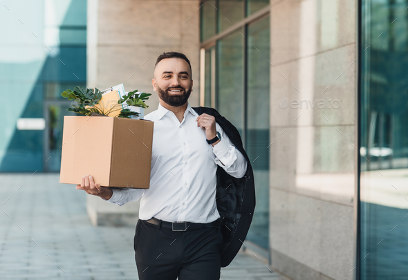 Happy middle-aged businessman walking outdoors with box of stuff as leaving business center, leaving