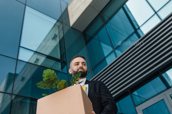 Upset mature businessman walking outdoors with box of stuff leaving business center, free space