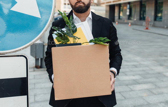 Unemployed businessman standing with box of stuff outdoors, near road sign, lost his job, crop