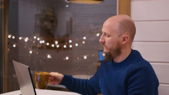 A Bald Man with a Beard Sits at a Table Working Remotely From Home After His Main Job in Order to