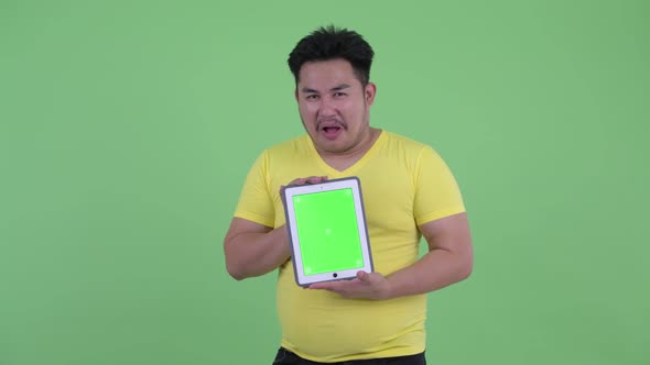 Happy Young Overweight Asian Man Showing Digital Tablet and Looking Surprised alt