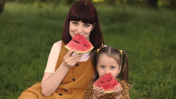 Young Mother Resting with Her Daughter in Park Touch the Noses and Holding a Watermelon alt