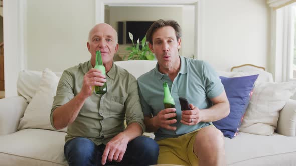 Portrait of happy caucasian grandfather with adult son with beer watching tv in living room alt