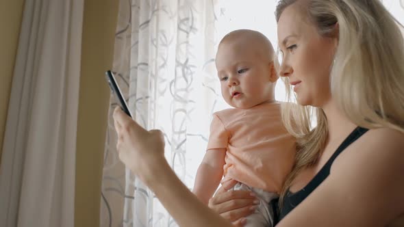 Young Mother Is Holding Baby, Looking at Smartphone Screen in Home Interior