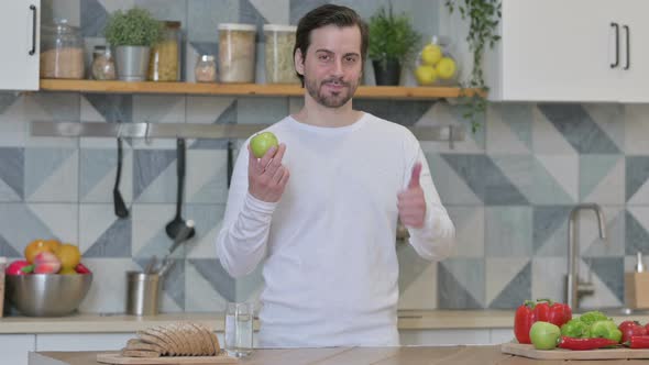 Young Man Showing Thumbs Up While Holding Apple in Kitchen alt