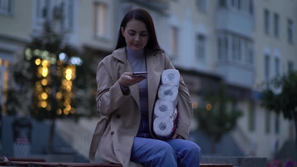 Woman Sits on Bench in Medical Mask with Toilet Paper and Smartphone During the Second Wave alt