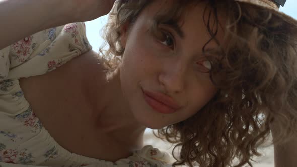 Portrait Curly Woman Playfully Adjusting Straw Hat on Head Relaxing on Seashore alt