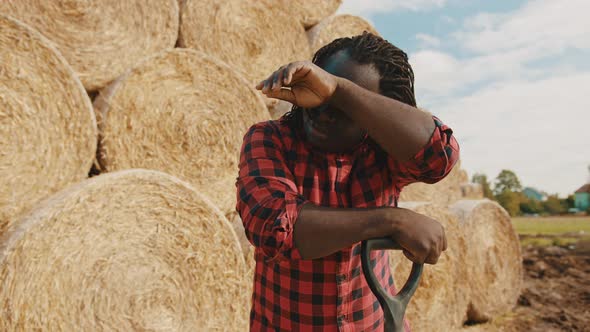 Young African Farmer Cleaning the Sweat with Hand From His Forehead While Resting on the Fork Handle alt