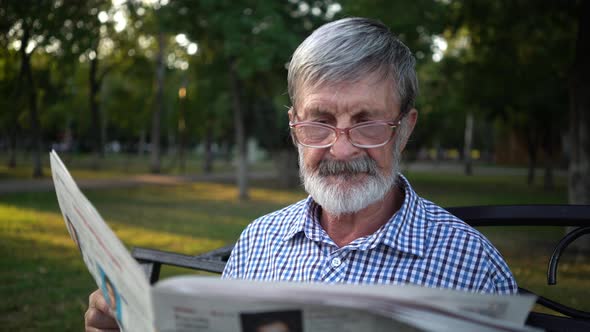 Senior in Plaid Shirt Sits on a Bench in the Park and Reads a Newspaper alt