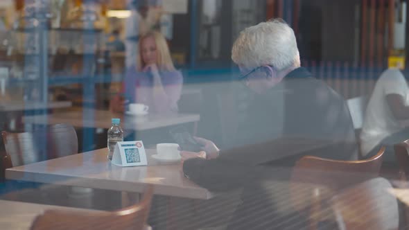 View Through Window If Mature Businessman Scan Qrcode with Cellphone in Restaurant alt