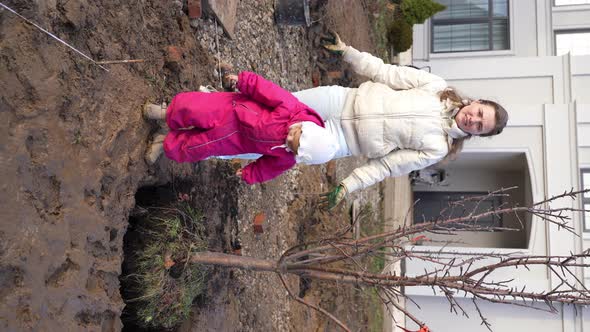 Happy Smiling Woman Plants Apple Tree with Her Little Daughter in Garden on Sunny Autumn Day alt