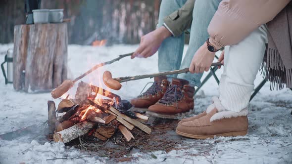 A Couple is Roasting Sausages on Sticks on a Campfire in the Woods in Winter