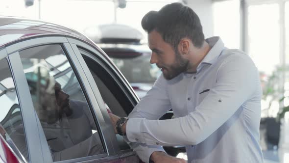 Handsome Man Smiling To the Camera While Examining Car for Sale at Dealership