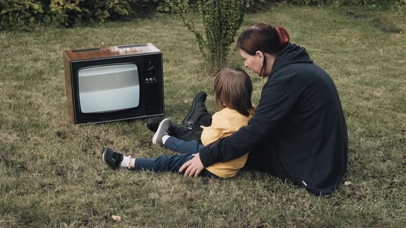 Woman Mother with Small Child are Sitting on Grass and Watching an Old Retro TV