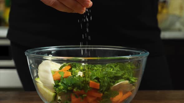 Closeup of a Woman's Hand Sprinkling Salt on a Salad in a Glass Bowl alt