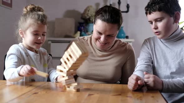 playful mom helps preschool children build tower of wooden blocks in living room