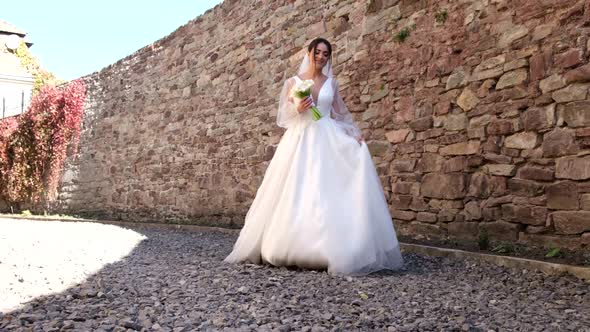 The Bride in a Luxurious Dress Walks and Laughs Against the Background of a Stone Wall alt