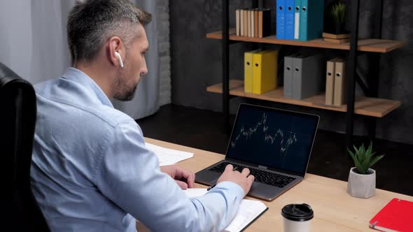 Man Trader Broker Makes Notes in Notebook Uses Laptop with Stock Exchange Chart alt