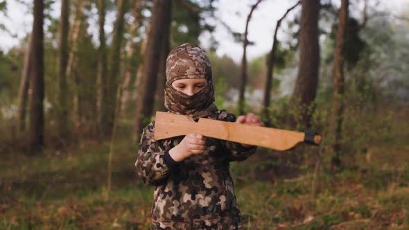 Little Boy in Camouflage with Wooden Toy Gun and in Balaclava Aims and Shoots Forward Then Reloads