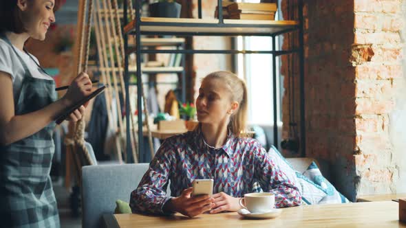 Pretty Girl Using Smartphone in Coffee House Ordering From Friendly Waitress alt