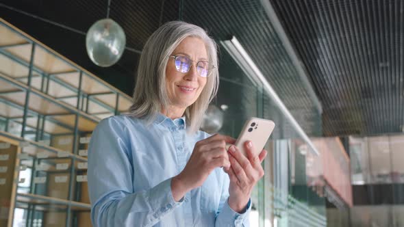 Grayhaired Senior Businesswoman is Using a Smartphone in an Office alt