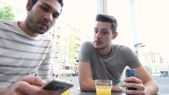 two young multi ethnic men outdoor sitting bar using smart phone having conversation alt