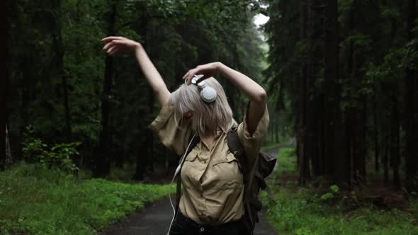 Dancing blond woman in headphones with backpack in rainy forest