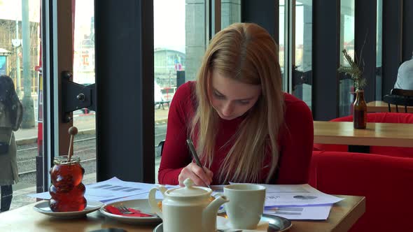 A Young Attractive Woman Sits at a Table with Meal in a Cafe, Looks Out a Window and Does Paperwork alt