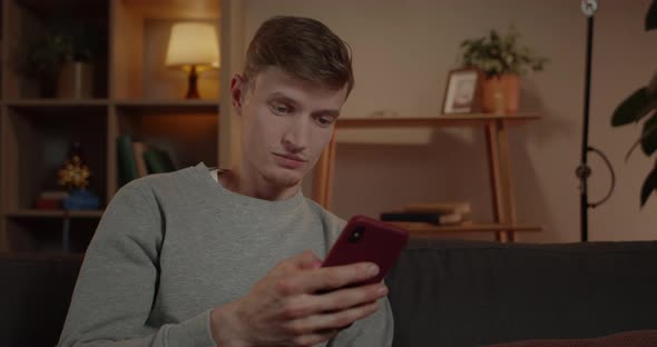 Crop View of Amazed Millennail Man Scrolling Phone Screen While Sitting on Sofa at Home alt