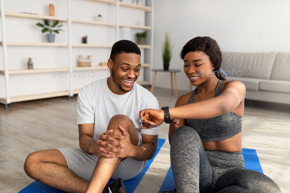Sporty black lady showing data on smartwatch or fitness tracker to her boyfriend after home workout - Stock Photo - Images