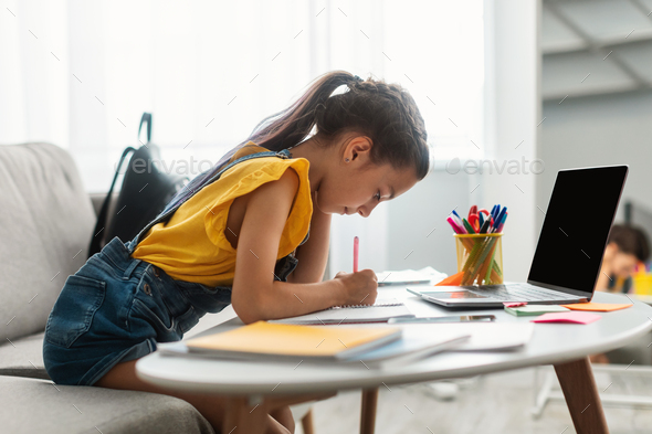 Girl sitting at desk, using blank laptop, writing in textbook