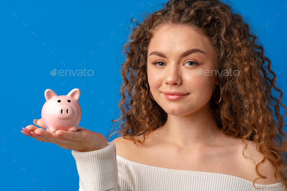 Curly-haired young woman holding piggy bank against blue background - Stock Photo - Images
