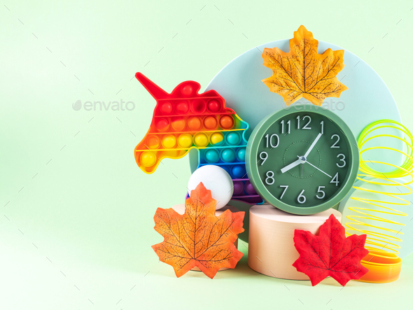 Hello autumn composition with orange leaves, geometric podiums, alarm clock and unicorn toy on green