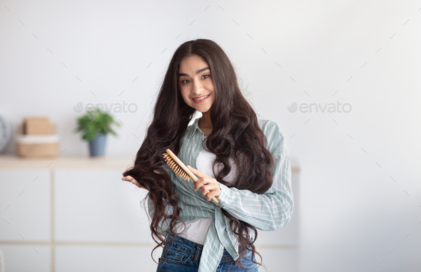 Portrait of cheerful Indian lady brushing her beautiful long hair, using wooden brush at home