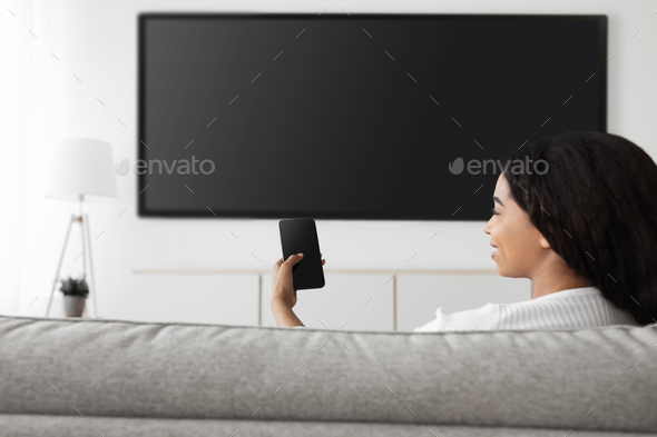 Smart home. Excited african american woman with cellphone connected to TV, plasma television with