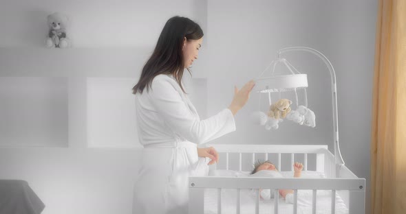 Mother in a White Coat Picks Up a Newborn Baby Lying in a Child Crib