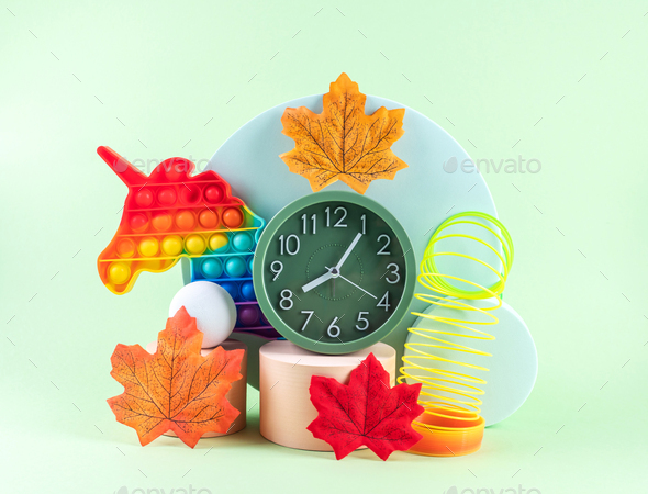 Hello autumn composition with orange leaves, geometric podiums, alarm clock and unicorn toy on green