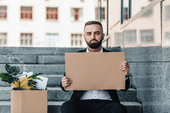 Sad middle-aged businessman sitting with empty poster and box of personal belongings on stairs near