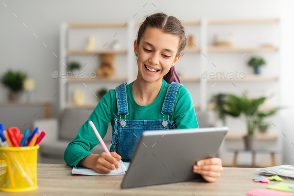 Girl sitting at table, using tablet, writing in notebook - Stock Photo - Images