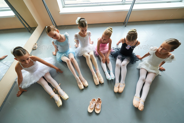 Six ballerinas sitting on floor, top view.