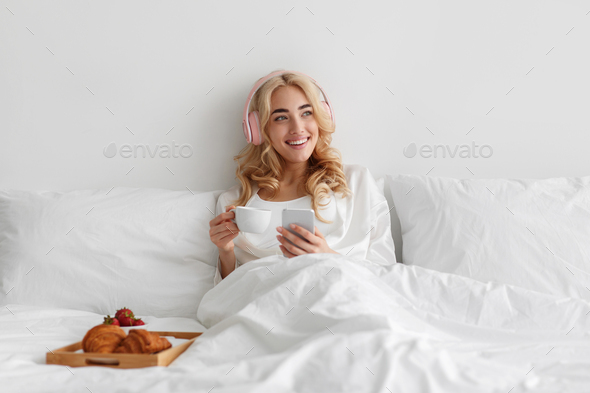 Young woman eats breakfast in bed listens music, enjoys free time, weekends and vacations