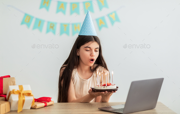 Indian teenage girl in festive hat having online birthday party, blowing candles on cake in front of